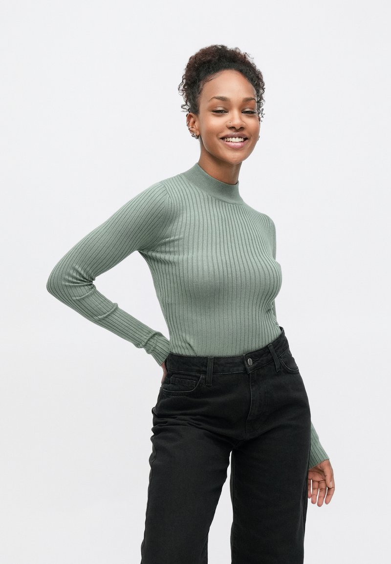 Ανοιχτό πράσινο ριπ τοπ με μακριά μανίκια και ψηλό λαιμό, σε συνδυασμό με μαύρο υψηλόμεσο τζιν με πλαϊνές τσέπες και λεπτή υφή.