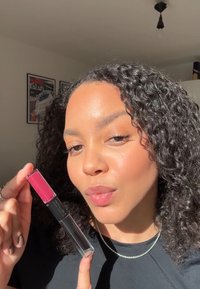 Rouge à lèvres dans un tube transparent avec un capuchon rose, présentant une texture crémeuse. Le modèle le montre sur un fond neutre avec de la lumière du soleil.