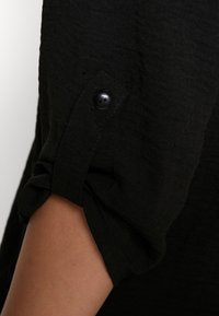 Sort tekstureret stof med en oprullet ærme, der har en sort knap. Nærbillede, der viser materialets overflade og detaljer.