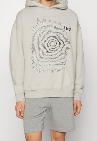 Sweat à capuche gris clair en tissu doux, présentant un motif en spirale noir sur le devant, avec une poche kangourou et des poignets côtelés. Porté avec un short gris.