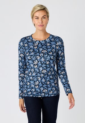 Vrouw die een navyblauw shirt met lange mouwen draagt met blauwe en beige bladpatronen, gecombineerd met een donkere broek, staand tegen een effen achtergrond.