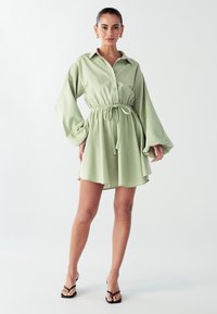 Šviesiai žalia, ilgomis rankovėmis suknelė marškiniai su mygtukų užsegimu priekyje, suspaustu juosmeniu ir laisva sijonu. Suderinta su juodais šlepetėmis.