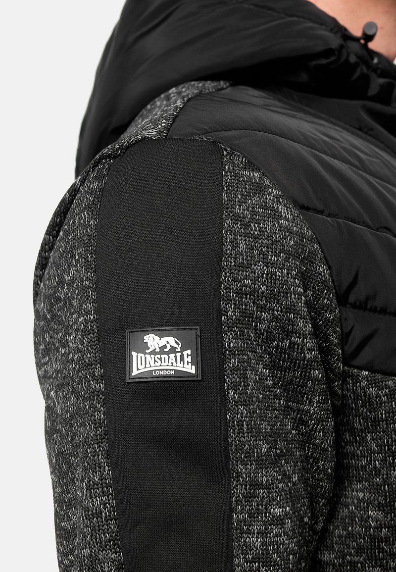 Zwart en grijs gewatteerd vest met textuur gebreide panelen, voorzien van een zwart stoffen schouder en een vierkant Lonsdale-logo op de linkerarm.