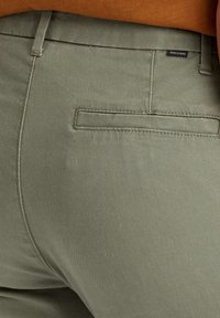 Olivgröna byxor med en slät yta, som har en bakficka och diskret sömnad. Etikett synlig på midjebandet.