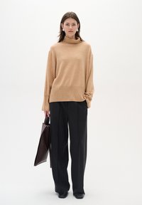Pulover bej cu guler înalt, cu croială lejeră, asortat cu pantaloni negri cu picior larg. Modelul ține o geantă tote maro închis. Fundal minim.