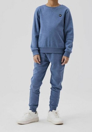 Ensemble sweat-shirt bleu et pantalon de jogging en tissu doux. Comprend un petit logo noir sur la poitrine et des baskets blanches. Coupe décontractée avec poignets côtelés.