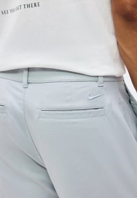 Pantalon Nike gris clair avec poches arrière passepoilées et logo Nike brodé, associé à une chemise blanche avec un texte noir partiellement visible.