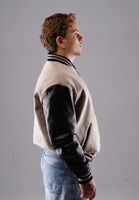 Beige Bomberjacke mit schwarzen Lederärmeln, Kragen mit horizontalem Streifen und Reißverschlusstaschen. Der Stoff ist glatt und hat eine strukturierte Passform.