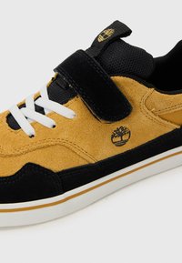 Baskets avec une tige en daim jaune, des accents noirs, des lacets blancs, une sangle ajustable et un logo sur le côté. Semelle en caoutchouc.