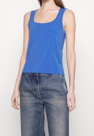 Kobieta ubrana w niebieski top na ramiączkach z kwadratowym dekoltem oraz wysokim stanem wyblakłych niebieskich dżinsów, stojąca na jednolitym tle.