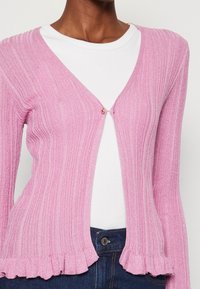 Lichtroze geribbelde cardigan met een V-hals, voorzien van een enkele gouden knoopsluiting en een gerimpelde zoom. Draagt over een wit shirt.