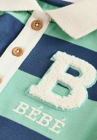 Polo cu dungi verzi și bleumarin, guler bej, butoane din lemn și litera "B" texturată în alb, cu "BÉBÉ" brodat dedesubt.
