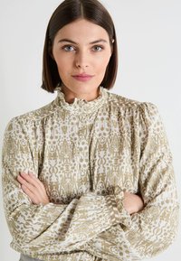 Blusa de color claro con un intrincado patrón marrón, cuello alto con volantes y mangas abullonadas con cierres de botón. Textura de tela suave.
