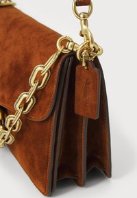 Brun ruskinds taske med guldfarvet hardware, en kædetræk og et lædermærke. Har en klaplukning og kontrastkant langs kanterne.