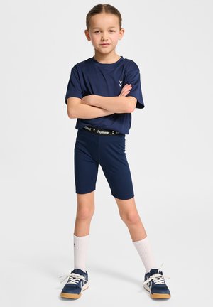 Marine sportoutfit bestaande uit een T-shirt met korte mouwen en fietsshorts. Voorzien van een wit logo en een zwarte elastische tailleband. Draagt met blauwe sneakers.