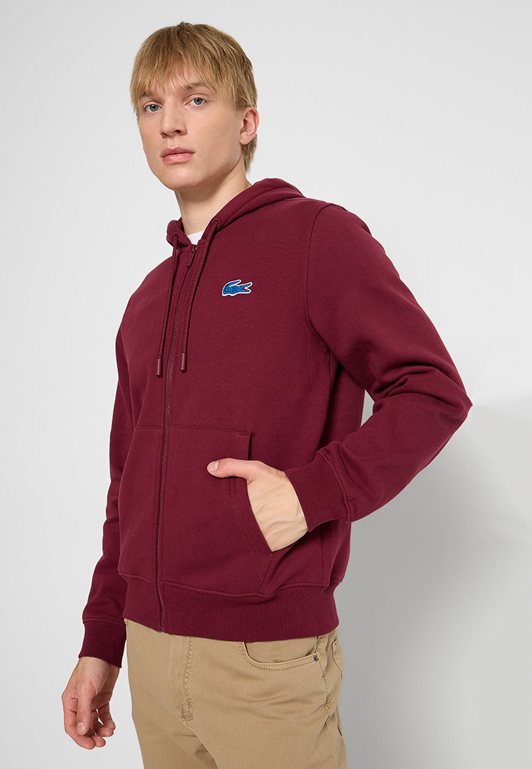 Sweat à capuche zippé bordeaux avec cordon de serrage, poche kangourou, poignets côtelés, et un logo Lacoste bleu sur la poitrine gauche. Texture de tissu lisse.