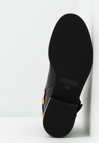 Semelle de chaussure noire vue de face avec la marque "See By Chloé" et la taille 37, montrant un motif de grip texturé et une partie de la tige noire ornée de clous dorés.