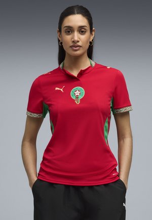Mujer con camiseta de fútbol marroquí roja con detalles verdes y logotipos dorados de Puma, de pie con las manos en los bolsillos de sus pantalones cortos negros, frente a un fondo gris.