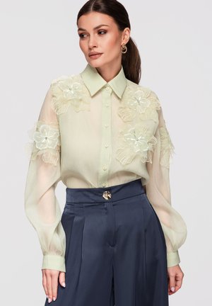 Frau trägt eine transparente hellgrüne Bluse mit großen Blumenapplikationen und goldenen Creolen, kombiniert mit hoch taillierten dunkelblauen Hosen.