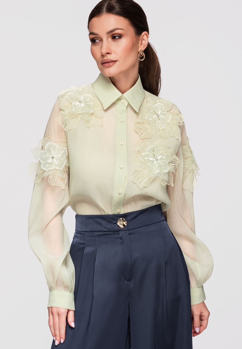 Femme porte un chemisier vert pâle transparent avec de grandes applications florales et des boucles d'oreilles créoles dorées, associé à un pantalon bleu marine taille haute.
