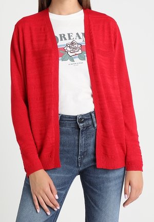 Kobieta w czerwonym otwartym kardiganie na białym t-shircie z różą i niebieskich dżinsach, ręce swobodnie wzdłuż ciała.