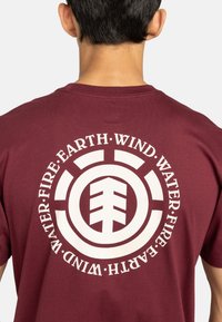 Camiseta de algodón color marrón con un gran gráfico en la parte posterior, que presenta un diseño circular y elementos de texto que representan Tierra, Viento, Fuego y Agua.