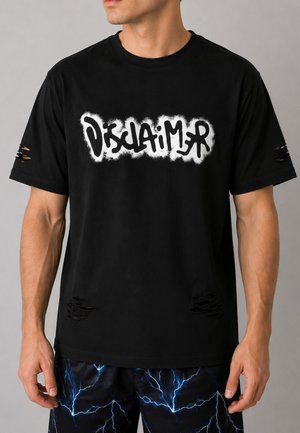 Uomo che indossa una maglietta nera consumata con scritta bianca in stile graffiti "Disclaimer" e pantaloncini neri con motivo a lampo blu.