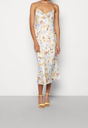 Mujer que lleva un vestido midi sin mangas con estampado floral, escote en V y tacones amarillos con tira en el tobillo, de pie frente a un fondo claro y sencillo.