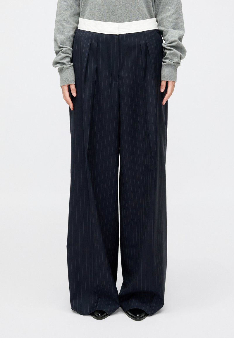 Tumši zila pinstripe platā līnija bikses ar baltu jostu, ar bārkstīm un gludu tekstūru, savienotas ar pelēku garam piedurkņu augšu.