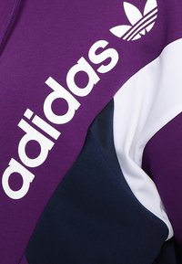 Giacca Adidas con una sezione superiore viola, accenti bianchi e blu navy, e un branding bianco prominente in un font audace e moderno.