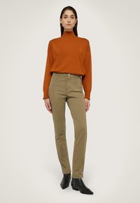 Verbrande oranje lange mouw top met een hoge hals, gecombineerd met lichtbruine slim-fit broek en zwarte enkellaarzen, staand tegen een neutrale achtergrond.