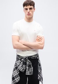 Νέος άνδρας με κοντά καστανά μαλλιά που φοράει ένα λευκό κοντομάνικο μπλουζάκι και ένα ασπρόμαυρο καρό πουκάμισο δεμένο γύρω από τη μέση του.