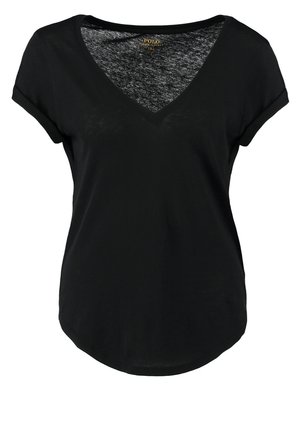 Kobieta idąca, ubrana w czarną bluzkę z dekoltem w serek, beżowe spodnie z brązowymi łatami na kolanach, czarne mokasyny i trzymająca brązową torebkę.