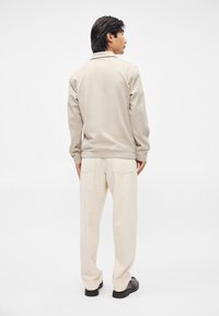 Jasnobeżowa kurtka o luźnym kroju, z widocznymi detalami szwów i elastycznymi mankietami, w połączeniu z kremowymi spodniami i czarnymi butami.