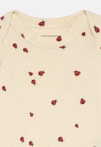 Baba obleka v kremni barvi z razpršenim vzorcem majhnih rdečih in črnih pikapolonic.