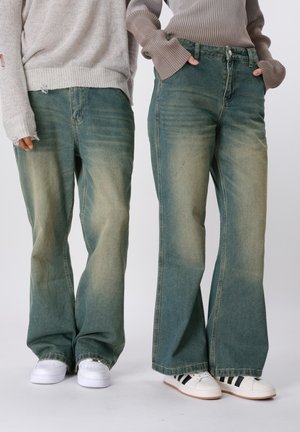 To personer iført falmede vidde jeans og afslappede sweatere, stående side om side med hænderne i eller nær lommerne.