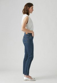Strakke blauwe denim jeans met een hoge taille, voorzien van een bruine leren patch en iconische achterzakstiksels. Gecombineerd met een witte mouwloze top.