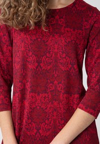 Czerwony top z długim rękawem z ozdobnym wzorem damaskowym, lekko teksturowaną tkaniną i okrągłym dekoltem oraz rękawami o długości do łokcia.