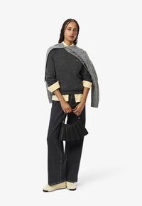 Maglione grigio chunky indossato sopra una camicia gialla chiaro, abbinato a jeans scuri e una borsa a pieghe nera. Scarpe bianche completano l'outfit.