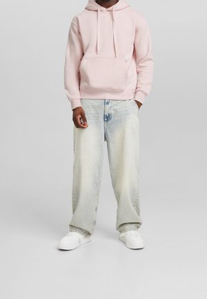 Felpa con cappuccio in cotone rosa chiaro con tasca frontale e coulisse, abbinata a jeans larghi azzurri e sneakers bianche. Outfit casual.