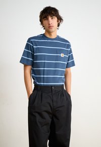 T-shirt rayé marine avec des lignes bleu clair, muni d'une poche poitrine et d'un patch logo ; porté avec un pantalon plissé noir. Tissu en coton lisse.