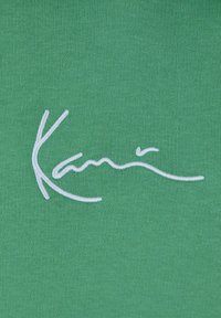 Tessuto verde con testo ricamato bianco che legge "Kari." La trama appare liscia con un motivo a maglia regolare.