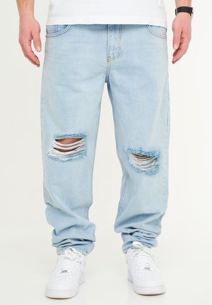 Light blue denim jeans with distressed rips at the knees, featuring frayed edges. Paired with white sneakers. Relaxed, wide fit.