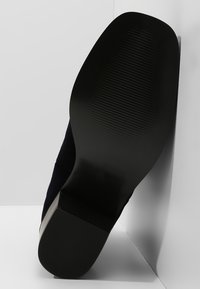 Suela de zapato negra con textura acanalada, vista en ángulo. La parte superior es de un material de ante azul marino profundo, que muestra un acabado suave.