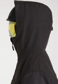 Personne portant une veste à capuche noire, un casque noir et des lunettes de ski teintées de jaune, vue de profil sur un fond blanc.