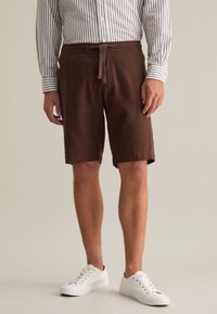 Shorts in lino marrone con vita elastica e fiocco anteriore, dotati di tasche laterali e una vestibilità rilassata. Abbinati a sneakers bianche.
