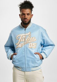 Veste bomber bleu clair avec des poignets et un ourlet côtelés, arborant un motif de logo crème et une fermeture éclair à l'avant. Tissu lisse et brillant avec une coupe décontractée.