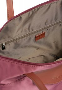 Interno di una borsa a tracolla in tessuto rosa con rivestimento beige, dotata di una tasca con zip e un'etichetta in pelle marrone. Texture liscia e design minimalista.