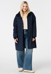 Marineblauwe waterdichte jas met een zwarte fleece-gevoerde kraag, drukknoopsluiting en zijzakken, gedragen over een lichte trui en wijde jeans.