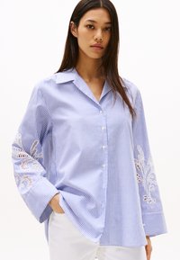 Camicia a righe blu e bianche con maniche lunghe e un dettaglio floreale in pizzo sulla manica sinistra; presenta un colletto classico e chiusura con bottoni.